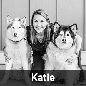 A woman is crouching between two huskies, smiling at the camera. The photo is in black and white, with a name tag at the bottom that reads "Katie." The background consists of wooden paneling.