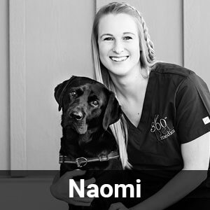 Black and white photo of a smiling person with long hair in a braid, wearing a scrub top with "360°" embroidered, sitting next to a black Labrador retriever. Both appear content. The name "Naomi" is written at the bottom of the image.