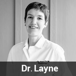 Black and white photo of a person with short hair dressed in a lab coat, smiling at the camera. The text "Dr. Layne" is written at the bottom of the image.