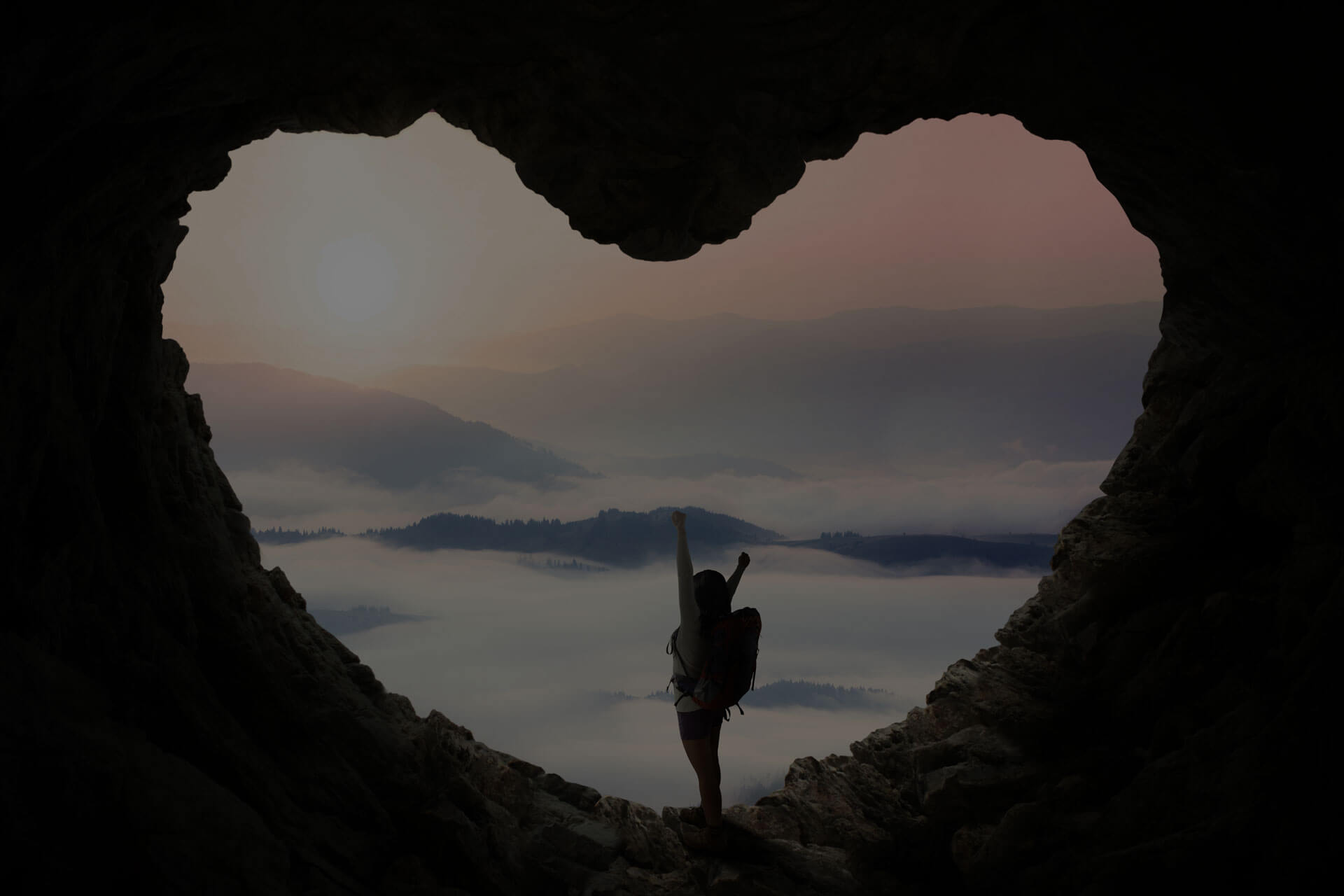 A person with a backpack stands at the opening of a heart-shaped cave, facing misty mountains and a foggy landscape under a gently colored sky at sunset or sunrise. The scene evokes a sense of adventure and awe.