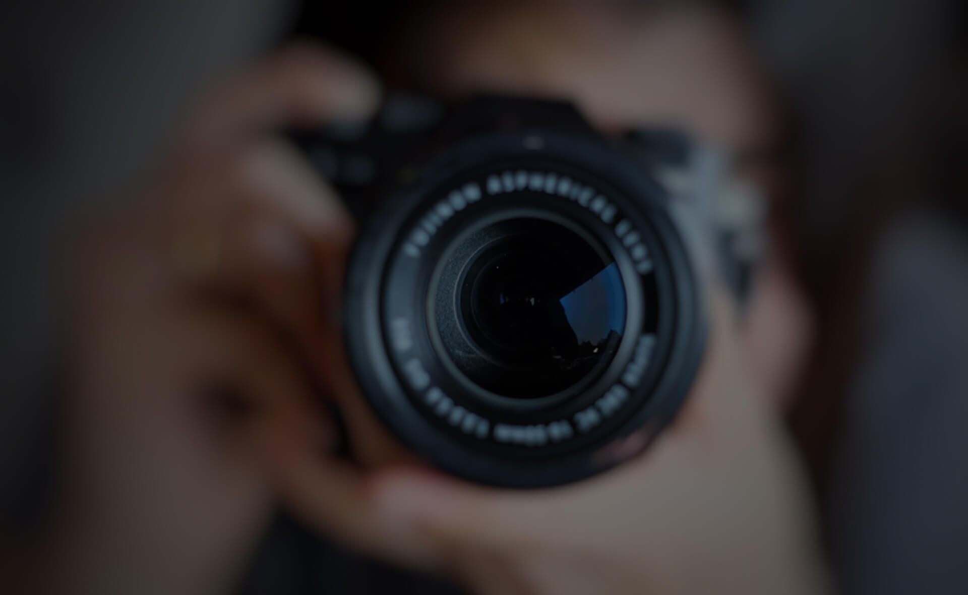 A close-up shot of a person holding and pointing a camera directly at the viewer. The camera lens is prominently featured and in sharp focus, while the person's face and background are blurred.