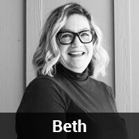 Black and white photo of a person with wavy hair and glasses, smiling at the camera. They are wearing a dark turtleneck and standing in front of a wooden wall. The name "Beth" is displayed in white text on a black strip at the bottom of the image.