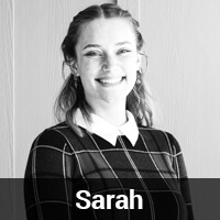 Black and white photo of a smiling woman with medium-length hair. She is wearing a checkered top with a white collar and has earrings. There is a label at the bottom with the name "Sarah.