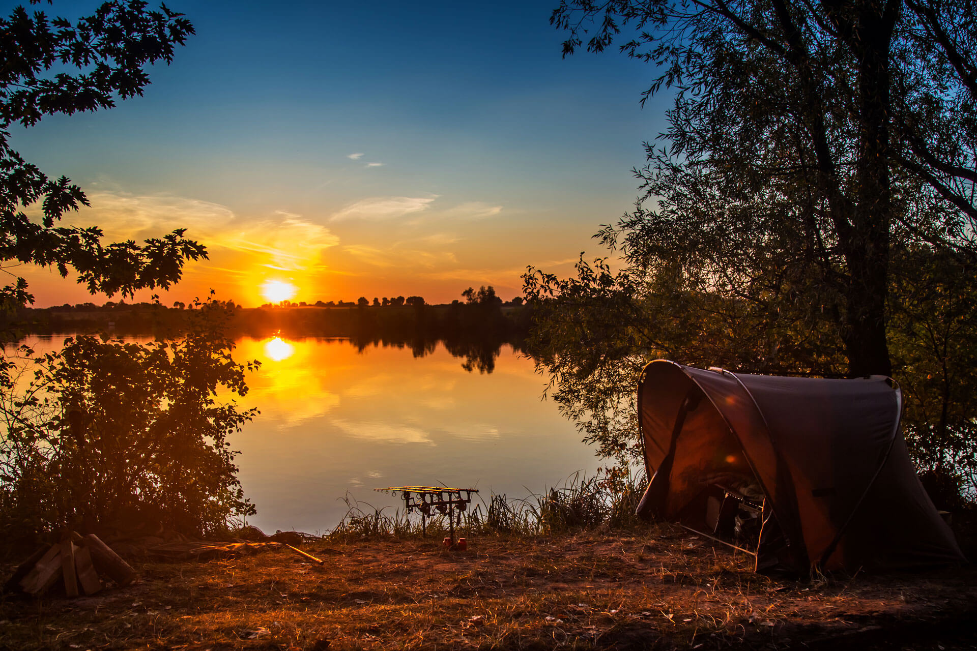 A serene lakeside scene at sunset with vibrant yellow and orange hues reflecting off the water. A camping tent is set up on the right side near the water's edge, surrounded by trees and foliage, with a small campfire wood stack visible on the left.