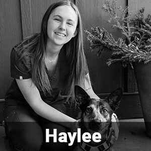 A young woman, identified as Haylee, is kneeling next to a dog in this black-and-white image. She is smiling and has one arm around the dog, which is sitting attentively. There is a potted plant visible in the background.