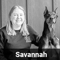 A smiling person with shoulder-length hair wearing a dark sweatshirt sits next to a large, alert Doberman. The bottom of the image has the name "Savannah" in white text on a black background.