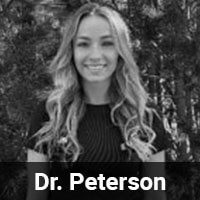 Black and white photo of a smiling woman with wavy hair, wearing a dark top, standing outdoors in front of trees. Text at the bottom reads "Dr. Peterson.